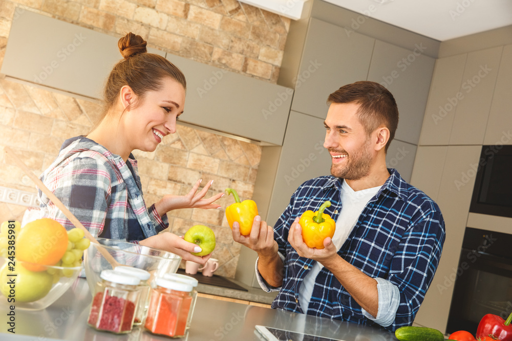 Wall mural young couple at home standing in kitchen together playing with vegetables cheerful