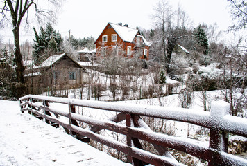 Wooden bridge in winter, trees and and wooden houses covered by snow in a village