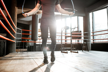 Young athletic man in black uniform training with a jumping rope, warming up on the boxing ring in...
