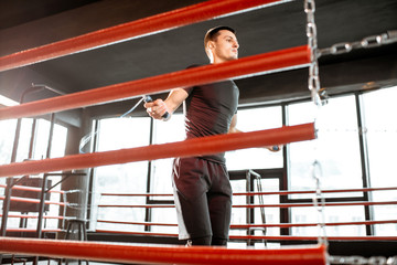 Young athletic man in black uniform training with a jumping rope, warming up on the boxing ring in the gym