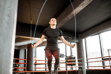 Young athletic man in black uniform training with a jumping rope, warming up on the boxing ring in...