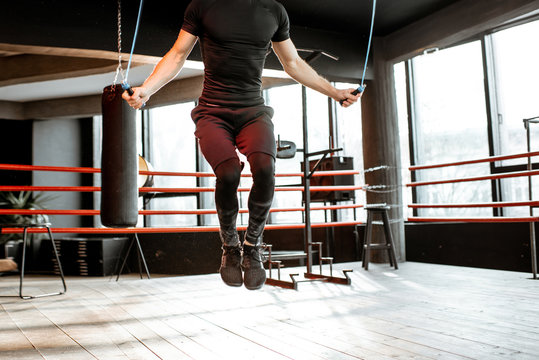 Young athletic man in black uniform training with a jumping rope, warming up on the boxing ring in the gym. Croppped image with no face