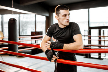 Fototapeta na wymiar Portrait of a young athletic man winding bandage on the wrists, preparing for training on the boxing ring at the gym