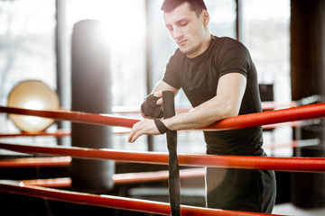 Portrait of a young athletic man winding bandage on the wrists, preparing for training on the boxing ring at the gym