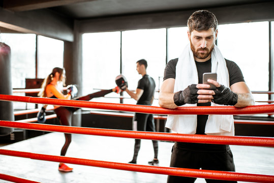 Man resting with phone after the training on the boxing ring with people boxing on the background at the gym - Powered by Adobe