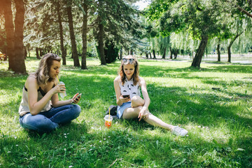 Two hipster girls are sitting on the grass with summer cocktails. Sisters are looking at their smartphones. Social networks communication. Cold non-alcoholic drinks with ice to go. Mojito and lemonade