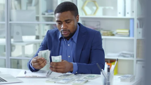 African american businessman sitting at desk with dollar stacks on it, counting money and writing down on document while working in the office