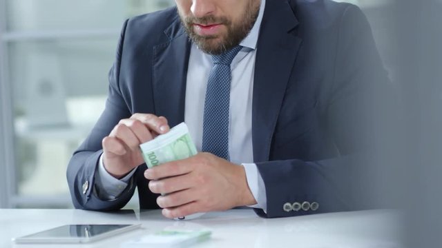 Mid-section of businessman counting euro banknotes and tapping on digital tablet while working at desk in the office