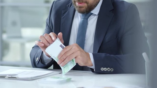 Mid-section of bearded businessman in formal suit counting euro banknotes and writing down on clipboard while working at desk in the office