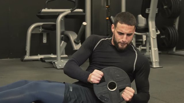 Athletic handsome young man with beard in black sportswear swinging press in gym, holding barbell plate in hands. Medium shot. Working out concept.