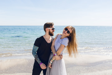 Guy with tattoo on hand is hugging girl with long hair near sea. He is looking at her. She is smiling to the camera.
