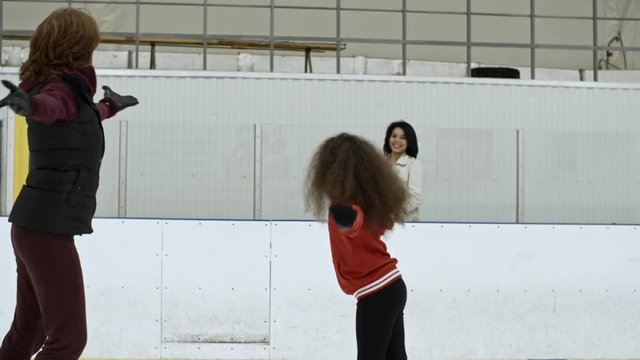 Little girl and female coach performing figure skating element on ice while mother watching them; daughter skating towards edge of rink and embracing mom
