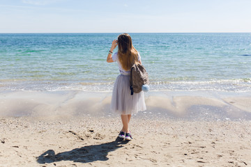 Young girl with long hair  and bag on back is looking to the sea. She wears lush gray skirt and...