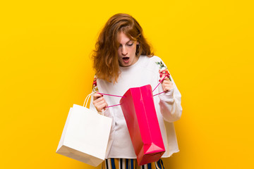 Young redhead woman over yellow wall surprised while holding a lot of shopping bags