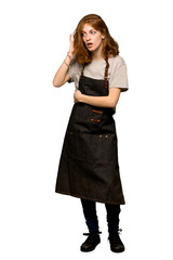 A full-length shot of a Young redhead woman with apron listening to something by putting hand on the ear over isolated white background