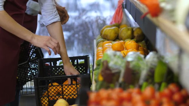 Close-up of multi ethnic grocery workers hands in aprons arranging organic tangerines and pears on store shelves. Shop clerks filling up storage racks in fruit and vegetable department of supermarket