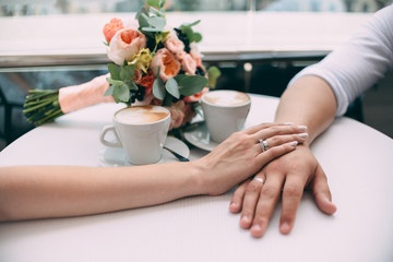 The bride and groom hold hands, holding expensive wedding rings with white gold, on the background of a wedding bouquet and two cups of coffee on a white table.