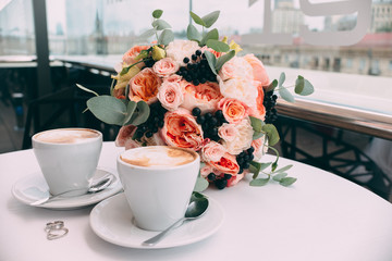 wedding rings on a white table on the background of a wedding bouquet and two cups of coffee.