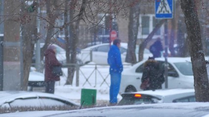 Man in blue waiting watching traffic and people on city street intersection during snowfall. Slow motion.