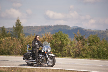 Side view of bearded biker in sunglasses and black leather clothing riding cruiser motorbike along narrow asphalt path on sunny summer day