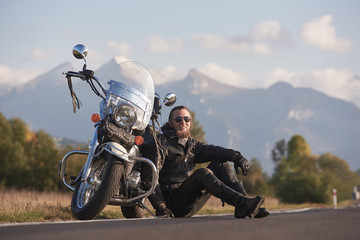 Smiling handsome motorcyclist in black leather clothing sitting at modern powerful shiny motorbike on grassy roadside on blurred copy space background of distant mountain peaks under bright sky.
