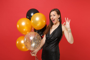 Fototapeta premium Amazed young girl in little black dress celebrating, showing OK sign, holding air balloons isolated on red background. International Women's Day, Happy New Year, birthday mockup holiday party concept.