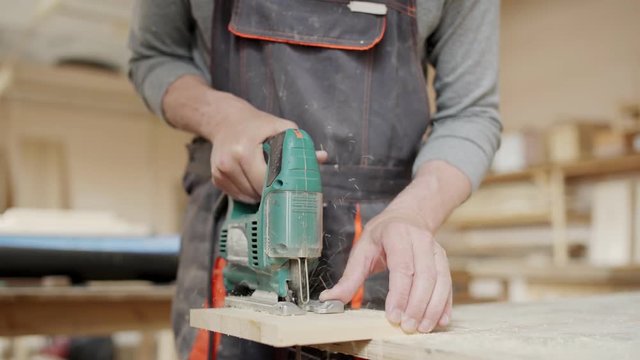Hands of carpenter using speed jig saw to cut wooden plank attached to workbench and then unscrewing vise
