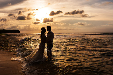 Silhouettes of the bride and groom look at each other standing in the sea water on the beach at sunset