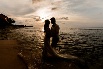 Silhouettes of bride and groom embracing standing in the sea water on the beach at sunset