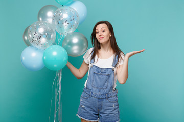 Bewildered young woman in denim clothes spreading pointing hand aside celebrating, holding colorful air balloons isolated on blue turquoise background. Birthday holiday party, people emotions concept.