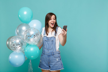 Fototapeta premium Surprised young woman in denim clothes looking on mobile phone celebrating, holding colorful air balloons isolated on blue turquoise wall background. Birthday holiday party, people emotions concept.
