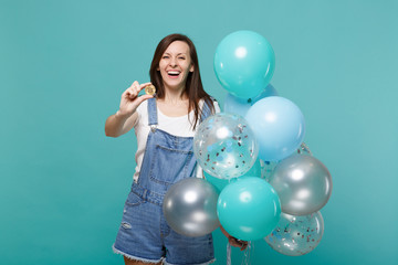 Fototapeta premium Laughing young woman in denim clothes holding bitcoin, future currency, celebrating with colorful air balloons isolated on blue turquoise background. Birthday holiday party, people emotions concept.