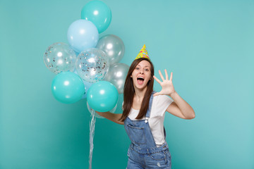 Funny young woman in birthday hat keeping mouth open showing palm and celebrating, holding colorful air balloons isolated on blue turquoise background. Birthday holiday party, people emotions concept.