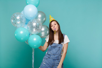 Pretty young woman in denim clothes, birthday hat blowing in pipe, celebrating and holding colorful air balloons isolated on blue turquoise background. Birthday holiday party, people emotions concept.