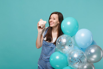 Fototapeta premium Portrait of pretty young woman in denim clothes holding piggy money bank, celebrating with colorful air balloons isolated on blue turquoise background. Birthday holiday party, people emotions concept.