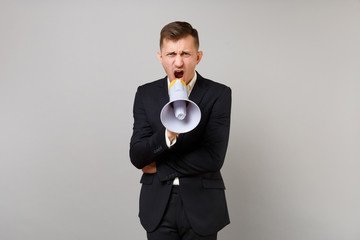 Portrait of nervous young business man in classic black suit, shirt screaming on megaphone isolated on grey wall background in studio. Achievement career wealth business concept. Mock up copy space.