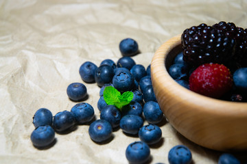 An isolated wooden bowl full of blueberries and wildberries with plain soft background decorated by fresh mint and with a few blueberries around on the paper