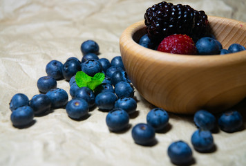 An isolated wooden bowl full of blueberries and wildberries with plain soft background decorated by fresh mint and with a few blueberries around on the paper