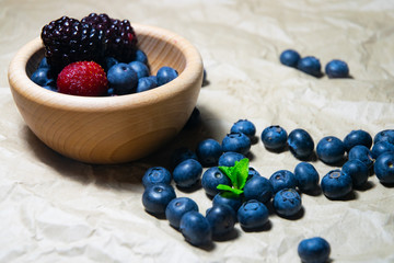 An isolated wooden bowl full of blueberries and wildberries with plain soft background decorated by fresh mint and with a few blueberries around on the paper