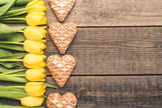 Beautiful yellow tulips and handmade gingerbread hearts create a frame with free, copy space. Lovely flowers and sweets lie on old wooden and natural boards. Fresh and sweet ingredients for lovers.