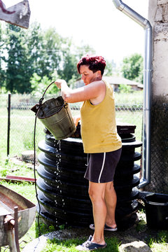 Full Body Portrait Of A Woman Fetching Water From A Natural Well. The Farmer Working Hard Filling Up The Shallow Basin. Daily Household Chore. Rural Area Scene