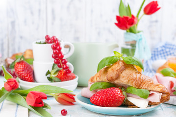 Sweet spring breakfast with croissants and bacon on a light background. A bouquet of red tulips and fresh berries of strawberries and currants. The concept of holiday celebration.