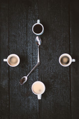 Coffee cups aranged as a clock face on a wooden black background. Four espresso mugs like a symbol of time with two spoons. Time for coffee. Breakfast time. Good morning concept.
