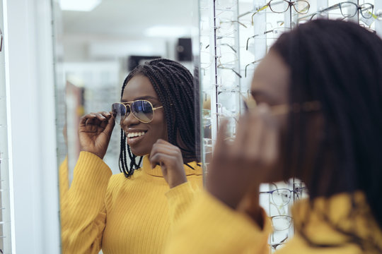 Young pretty african woman in yellow sweater choose sunglasses in Ootician's shop