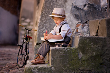 A little boy, dressed in a retro style, sits on the stairs of the old town, draws, next to it stands a bicycle. The historic street of the Polish city of Lublin.