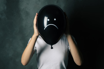 Woman hiding behind black balloon with drawn sad face on dark background. Depression symptoms