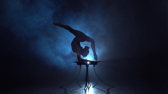 Acrobat on the table shows the tricks of standing on her hands . Smoke background. Silhouette