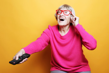 lifestyle and people concept: funny grandmother is holding a TV remote over yellow background 