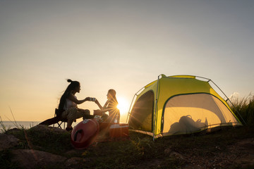 Two asian female hikers sit and clinking glasses by tent.