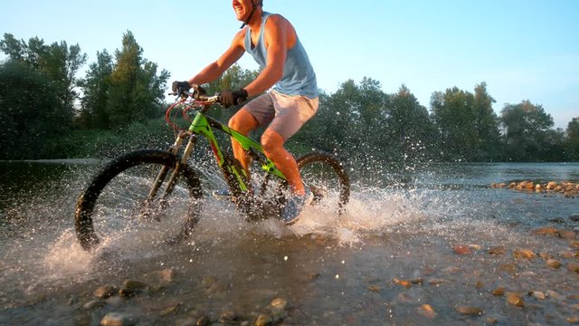 SLOW MOTION TIME WARP: Happy Caucasian mountain biker riding in the shallow river water on a sunny summer evening. Active man splashing the glassy stream water as he speeds downhill on mountain bike.
